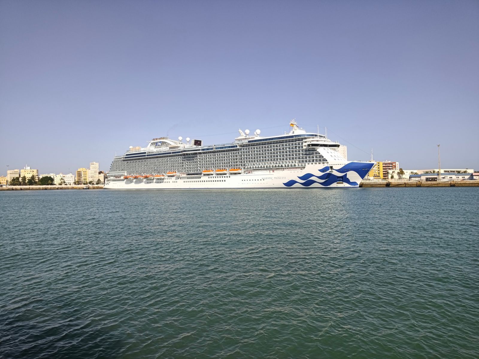 A large cruise ship berthed across a calm bay, rows of balcony and oceanview cabins along its side