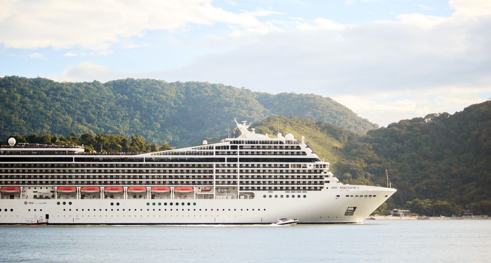 A cruise ship sailing past green hills in calm water, the kind of trip cruise savings can pay for